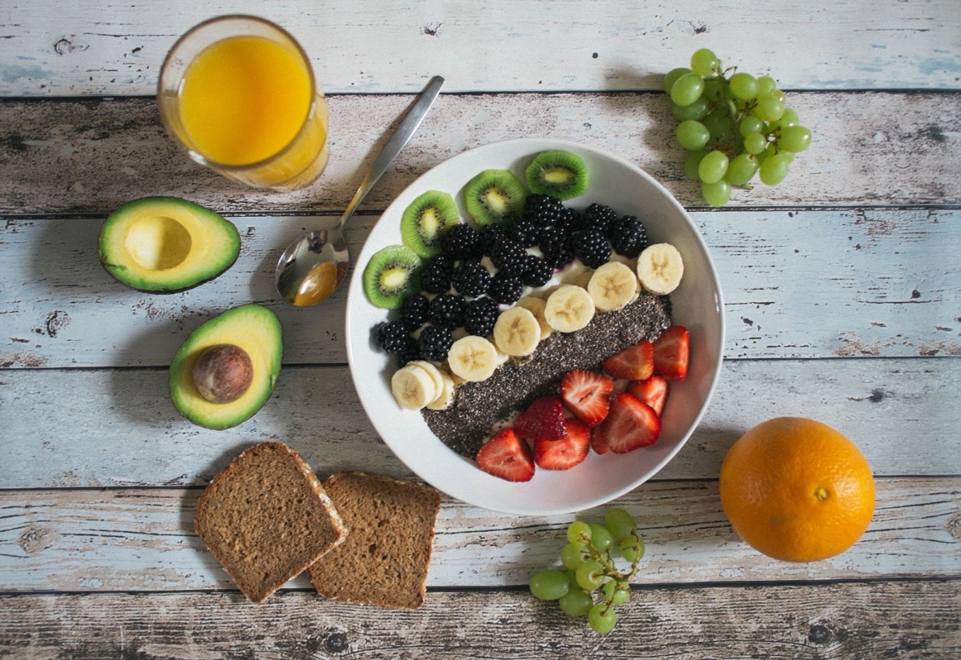 A bowl of fruits and chia seeds with other fruits, bread and juices besides it.